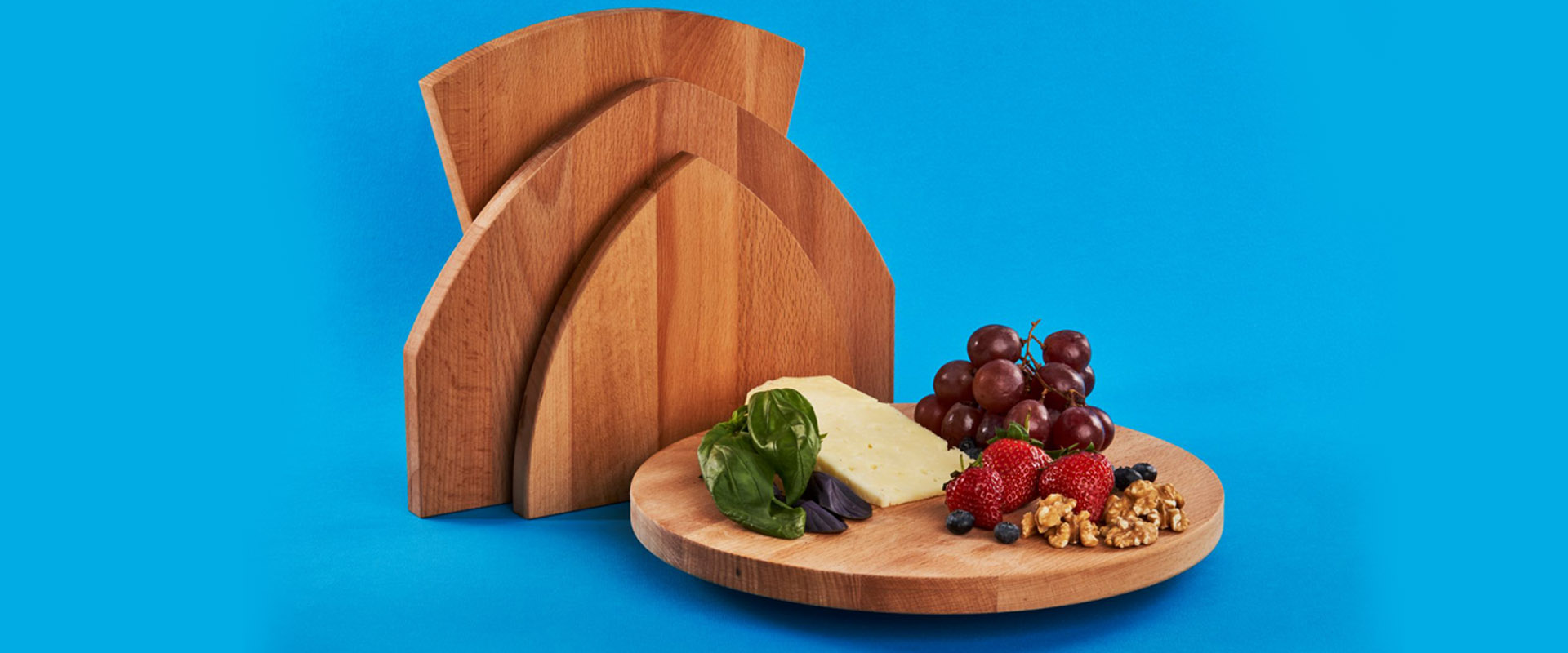 Wooden cutting boards of varying shapes displayed with cheese, grapes, berries and herbs against a bright blue background