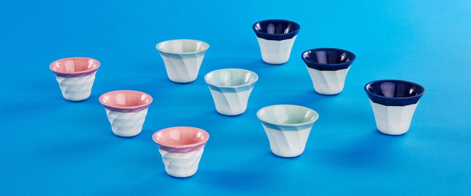 A set of eight small ceramic cups with colorful pink, blue and navy interiors arranged on a bright blue background