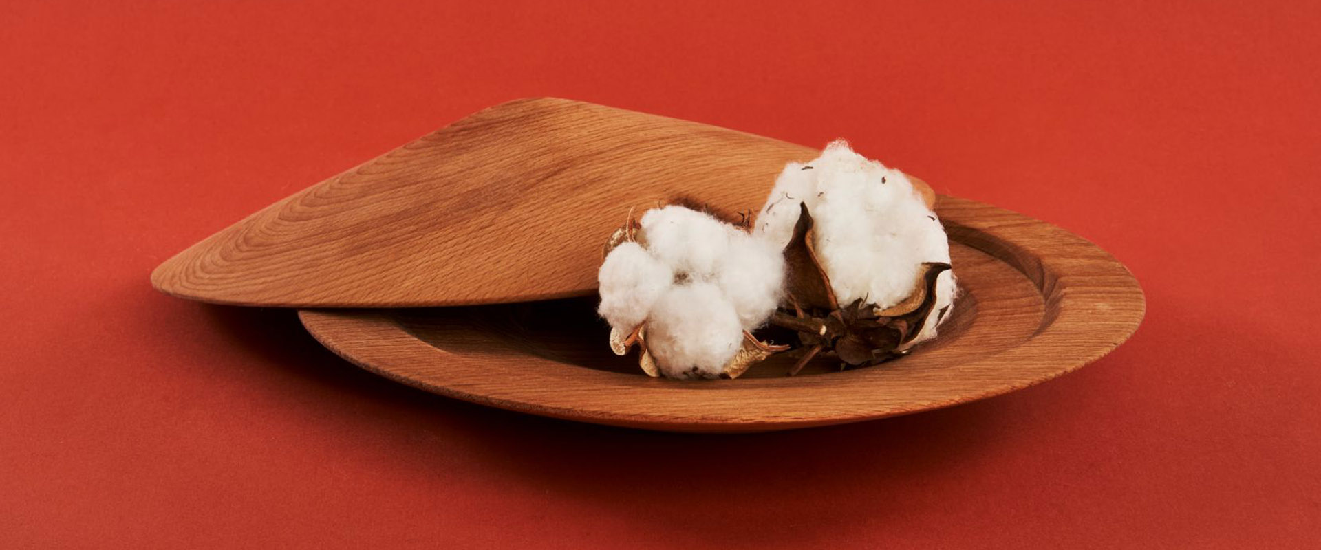 A wooden bowl with a hinged lid partially open, revealing cotton bolls inside, against a rich red background