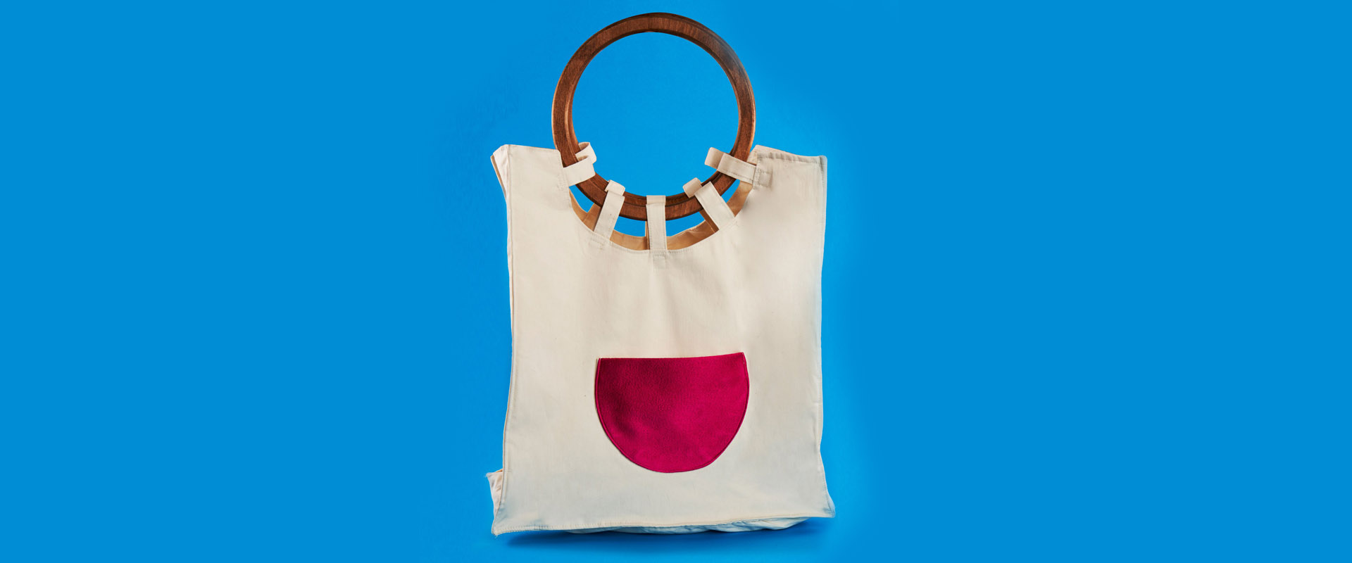 A cream canvas tote bag with a round wooden handle and a pink semicircular pocket, against a blue background